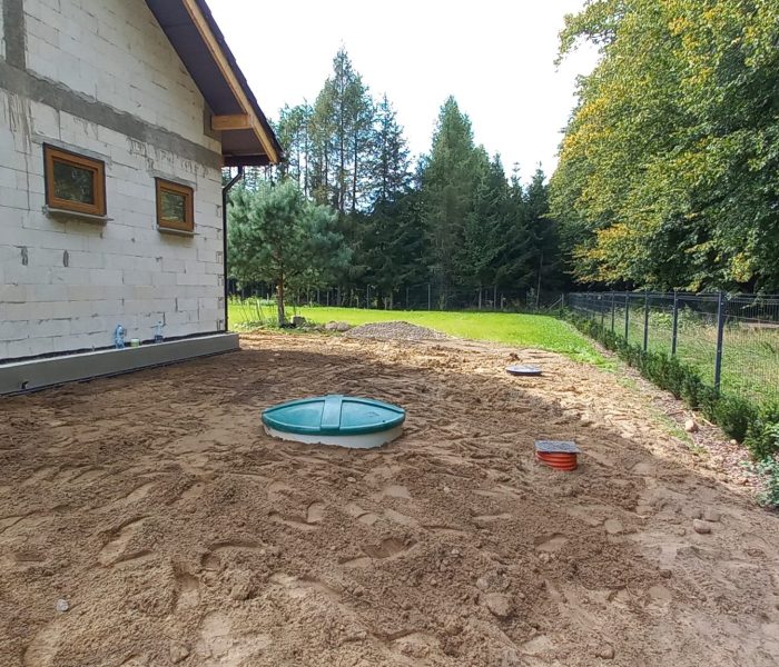 Budynek z białymi ścianami i podwójnymi oknami. Obok niego ogródek z siatką, drzewami i zielonym trawnikiem. Przy domu pok...